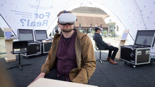 Zwei Männer sitzen während des Auftakts des VR-Projekts "In Echt? Virtuelle Begegnung mit NS-Zeitzeuginnen und Zeitzeugen" im Historischen Kutschstallhof in der mobilen Ausstellung. 