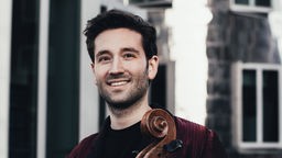 Der Cellist Roger Morelló Ros steht in einer Straße und hält sein Instrument.