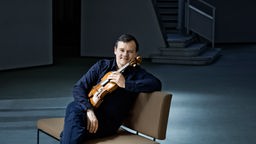 Der Geiger Frank-Peter Zimmermann hält sein Instrument in den Händen und sitzt auf einem modernen Sitzmöbel.