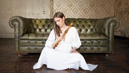 Die Blockflötistin Lucie Horsch sitz in einem weißen Kleid vor einem Sofa auf dem Boden.