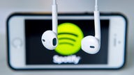 Kopfhörer hängen vor einem Apple Iphone 5s, auf dem das Logo vom Musik-Streaming-Dienst Spotify angezeigt wird.