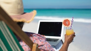 Frau mit Sonnenhut sitzt in einem Liegestuhl am Strand, sie hält ein Getränk in der Hand und hat ihren Laptop auf dem Schoß.