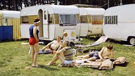 Mehrere Personen liegen und stehen auf einem Campingplatz neben vielen nebeneinander geparkten Wohnwagen