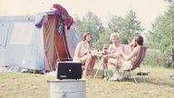 Eine Gruppe sitzt gemütlich essend an einem Tisch vor einem Zelt und hört dabei vermutlich Radio (1970er)