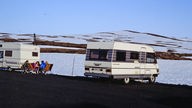 Zwei Wohnwagen stehen auf einem Campingplatz. Davor sitzen ein paar Urlauber zusammen und unterhalten sich. (1980er)