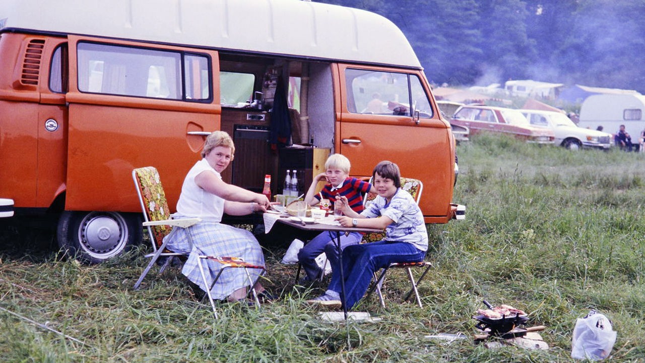 Eine Frau und zwei Kinder sitzen an einem Campingtisch am Bulli (1983)