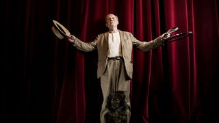 Ulrich Tukur steht bei einem Porträt-Termin mit seinem Hund Peppina 2022 vor einem roten Vorhang auf der Bühne und gestikuliert