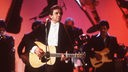 Johnny Cash (Sänger) bei einem Auftritt 1983 mit schwarzem Jackett und Fliege, spielt Gitarre, im Hintergrund Musiker