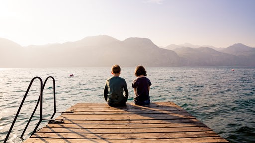 Zwei Kinder sitzen auf einem Steg an einem See und gucken aufs Wasser