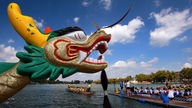 Ein langes Holzboot, auf dem Menschen sitzen. Eines der Boote befindet sich im Vordergrund des Bildes. Hier ragt der Kopf eines bunt bemalten Drachen ins Bild.