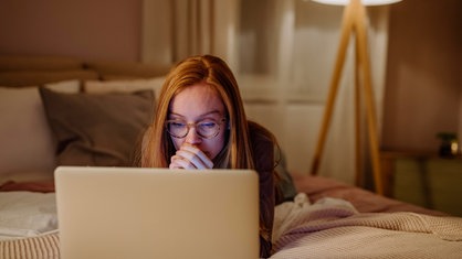 Eine Frau mit Brille liegt in einem Bett und schaut auf den Laptop