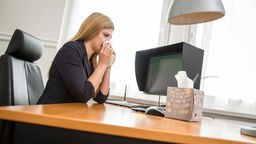 Eine Frau sitzt am Schreibtisch und putzt sich die Nase