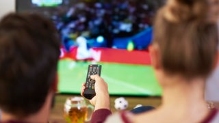 Zwei Menschen sitzen mit Fernbedienung in der Hand vor dem Fernseher und schauen Fußball