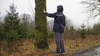 Ein Teilnehmer des Achtsamkeits-Spaziergang berührt mit der flachen Hand einen moosbewachsenen Baum