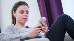 Eine junge Frau sitzt in einem Wohnzimmer und benutzt ihr Smartphone (gestellte Szene)