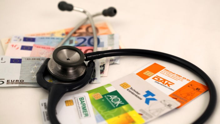Die Versichertenkarten der Krankenkassen DAK, AOK, Barmer und Techniker-Krankenkasse TK liegen mit Euro-Geldnoten unter einem Stethoskop