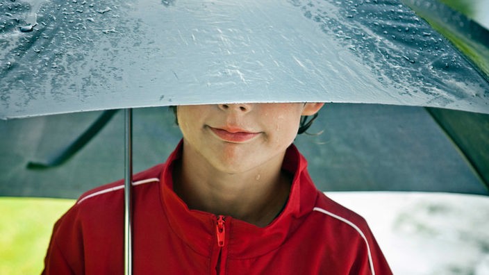 Ein Junge in roter Sportjacke lächelt unter einem nassen Regenschirm hervor, seine Augen sind verdeckt