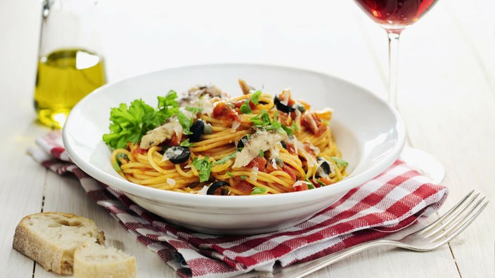 Ein weißer Porzelanteller, gefüllt mit Spaghetti in einer Oliven-Sardellen-Soße. Neben dem Teller steht ein Rotweinglas und eine Flasche Öl.