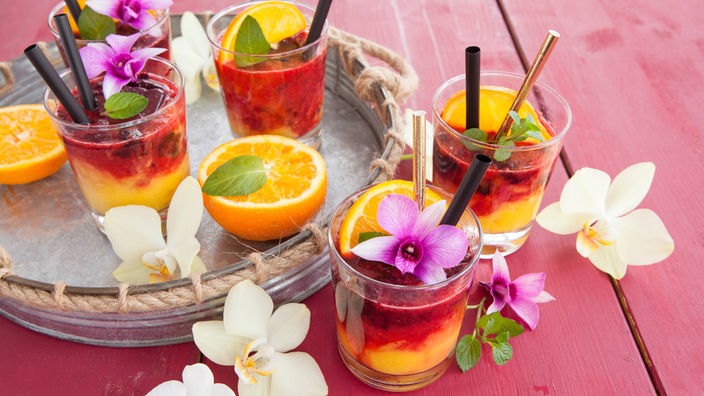 Bunte Cocktails auf Tablett mit Blüten und aufgeschnittenen Orangenhälften