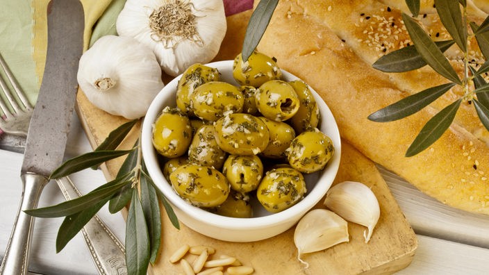Marinierte Oliven in einer kleinen Schale. Daneben Knoblauchzehen, Olivenbaumblätter und Fladenbrot.