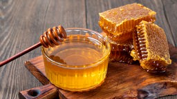 Eine Schale mit Honig steht auf einem hölzernen Brett. Dahinter befinden sich Honigwaben