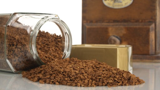 Das Granulat einer ausgekippten Dose Instant-Kaffee liegt auf einem Tisch vor einer Kaffeemühle.