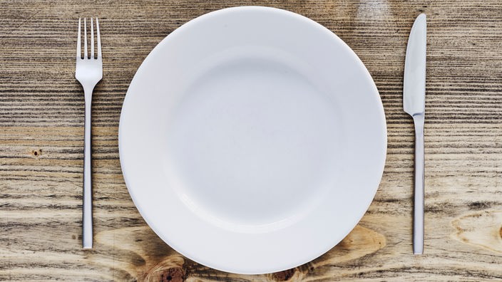 Ein leerer Teller steht auf einem Holztisch. Links eine Gabel, rechts ein Messer.