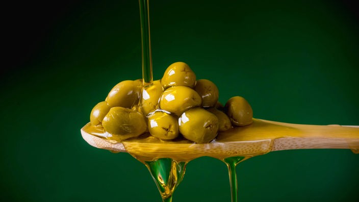 Nahaufnahme von Oliven auf einem Holzlöffel vor grünem Hintergrund. Ein Strahl Olivenöl wird darüber gegossen