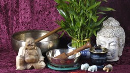 Klangschalen, Buddhastatuen und Räucherwerk vor einem purpurnen Tuch