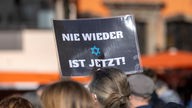 Ein Teilnehmer der Kundgebung "Aufstehen gegen Israelhass und Antisemitismus" hält ein Plakat mit der Aufschrift "Nie wieder ist jetzt" in die Höhe 