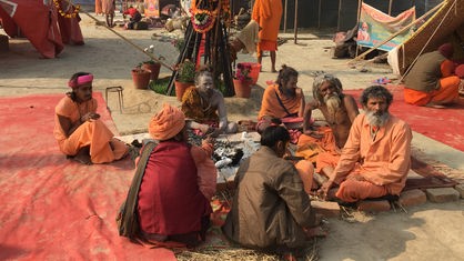 Sadhu Mönche in den typischen, orangen Gewändern sitzen im Kreis um eine Feuerstelle. Im Hintergund sind traditionelle Waffen zu sehen.