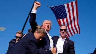 Donald Trump nach dem Anschlagsversuch am 13. Juli 2024 in Butler, Pennsylvania. Er wird von Personenschützern umringt. In seinem Gesicht sind Blutspritzer zu sehen, seine Faust ist gen Himmel gereckt. Im Hintergrund weht die US-Flagge. 