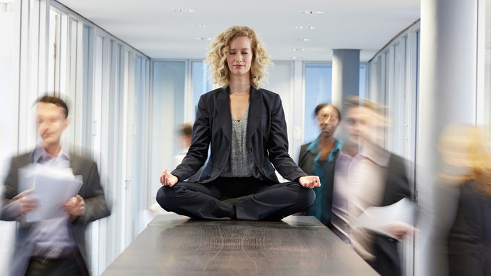 Eine Frau meditiert im Büro auf einem Tisch, um ihr herum geschäftige Menschen.