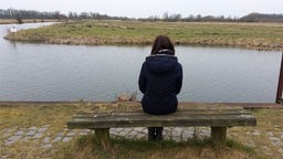 Eine Frau sitzt alleine auf einer Bank vor einem Gewässer-.