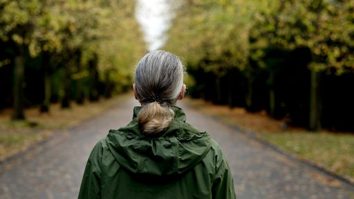 Das Beitragsbild des Dok5 Feature "Einsamkeit" zeigt eine ältere Frau mit grauen Harren von hinen alleine auf einer Baumallee stehen. 