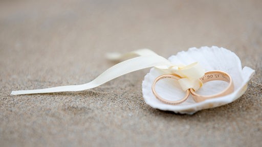 Das Beitragsbild zeigt zwei goldene Eheringe, verbunden mit einem weißen Seidenband in einer weißen Muschel am Sandstrand