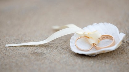 Das Beitragsbild zeigt zwei goldene Eheringe, verbunden mit einem weißen Seidenband in einer weißen Muschel am Sandstrand