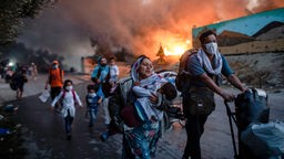 Ein brennendes Flüchtlingslager im Hintergrund. Davor fliehende Familien mit Kleinkindern. 