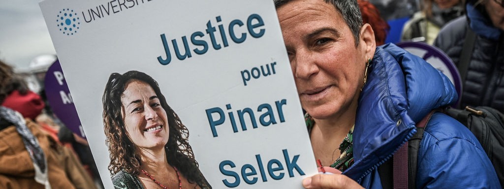 Das Beitragsbild des Dok5  "Pinar Selek – türkische Soziologin und Menschenrechtlerin" zeigt eine Demonstrantin vor dem Gerichtsgebäude in Istanbul 2023 mit einem Protestschild mit der Aufschrift Gerechtigkeit für Pinar Selek.  