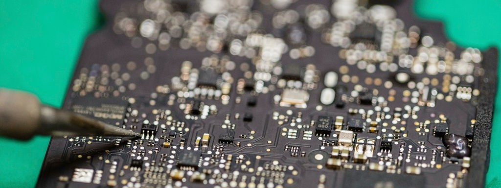 Beitragsbild zeigt die Nahaufnahme eines Laptop Microchips, der gerade mit Hilfe eines Lötkolbens repariert wird.