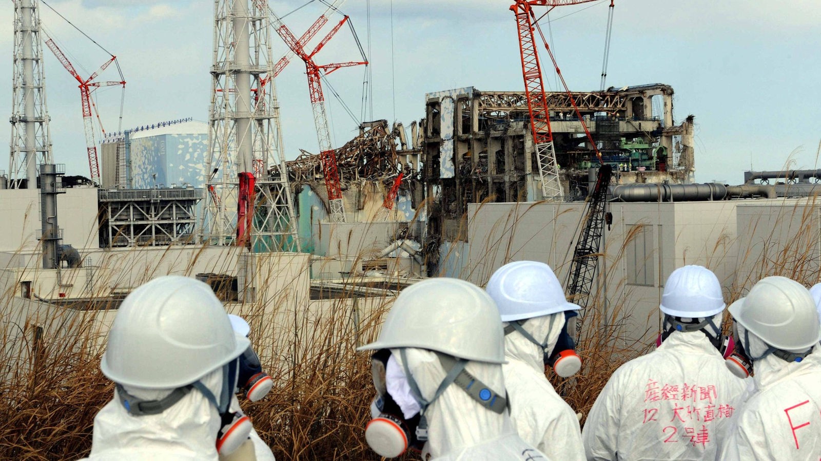 Das Beitragsbild des Dok5 Jenseits der Vorstellung zeigt Journalisten und Mitarbeiter von Tepco in Schutzausrüstung und Masken vor den Trümmern des Kernreaktors in Fukushima.