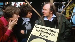 Karl Heinz Hansen bei einer Abrüstungsdemo 1981, er steht zwischen zwei Damen mit einem Schild um den Hals auf dem Steht "Für eine Atomwaffenfreie Zone"