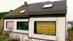 Ein Wohnhaus in der Dortmunder Neubau-Siedlung in Dorstfeld-Süd. Im Fenster ein Schild mit Schriftzug "Dorstfeld-Süd; Altlasten-Versuchsgebiet = Betrogene auf Gift" (1985).