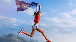 Mädchen springt mit einer EU-Flagge auf den Schultern vor einer Gebirgslandschaft. (Montage / Symbolbild gespiegelt) 