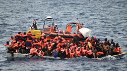 Ein Rettungsboot schwimmt hinter einem mit Migranten besetztem Boot im Mittelmeer. 