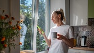 Eine Frau steht am Morgen entspannt in der Küche mit Kaffeetasse und Handy