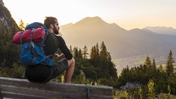 Ein junger Wanderer mit Rucksack macht in den Alpen Pause, sitzt auf einer Bank und blickt auf das Bergpanorama