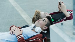 Zwei Jungs liegen mit ihren Köpfen auf einem Skateboard, sie haben ein Smartphone und Kopfhörer bei sich.