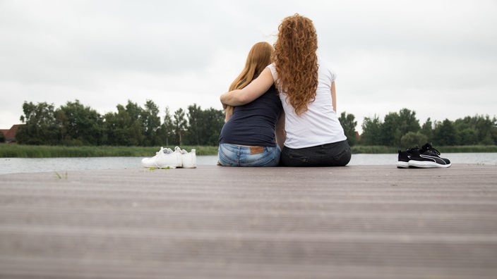 Symbolfoto: Zwei junge Frauen sitzen mit ihren Rücken zur Kamera gerichtet, auf einem Steg an einem See. Eine der Frauen hält die andere tröstend in ihrem Arm.