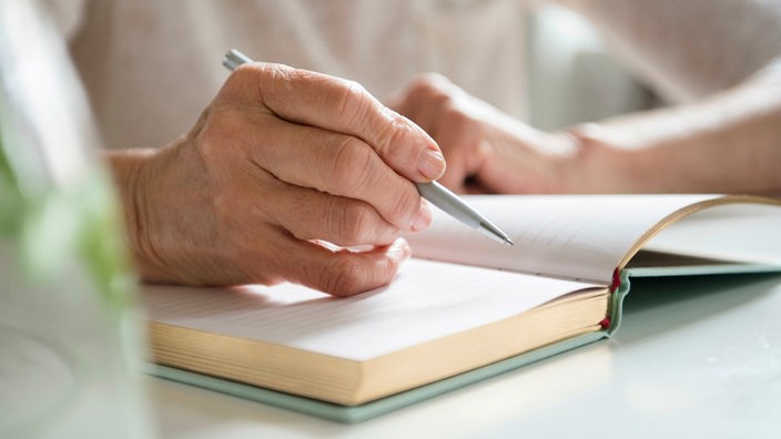 Symbolbild Intensivtagebuch: Das Bild zeigt die auf ein offenes Buch gelehnten Hände eines Menschen, der mit seiner rechten Hand einen Kugelschreiber hält.